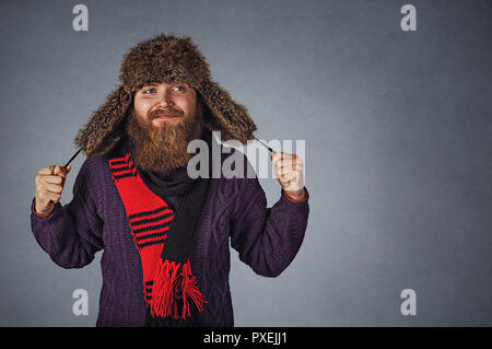 Closeup portrait stylish bearded man in russian fur hat cap and red scraf and dark purple sweater looking to side daydreaming thoughtful isolated on g Stock Photo