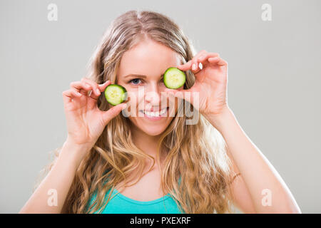 Beautiful young woman showing slices of cucumber on gray background. Stock Photo