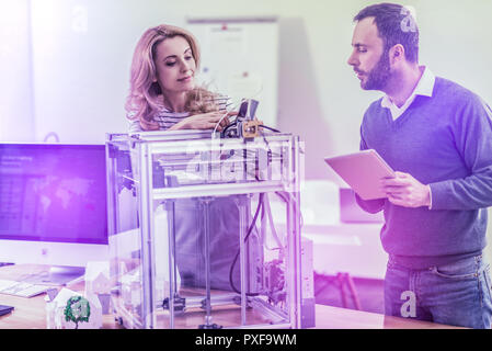 Blonde-haired beautiful officer worker fixing 3D printer standing near colleague Stock Photo
