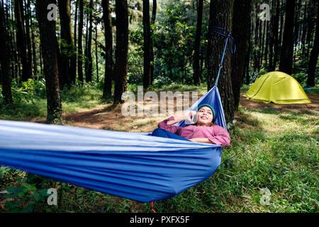 Smiling asian hiker woman talking on cellphone while lying in hammock on the forest Stock Photo