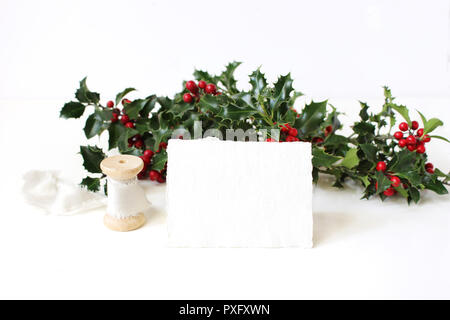 Festive Christmas mockup scene with handmade paper place card, spool of silk ribbon and holly red berries, leaves and branches on white table backgrou Stock Photo