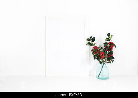 Vertical white blank wooden frame mockup. Holly berry branches in blue glass vase on white table. Styled stock feminine photography. Home decor. Christmas winter concept. Stock Photo