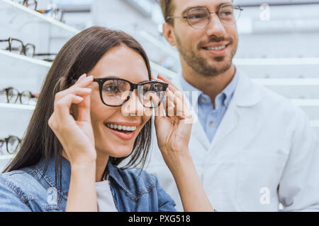 portrait of smiling woman choosing eyeglasses while male optometrist standing near in optics Stock Photo
