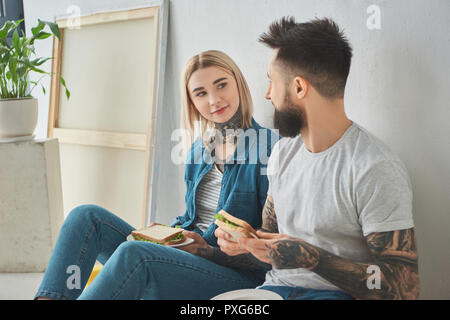 happy young couple holding sandwiches and smiling each other while sitting in floor in new house Stock Photo