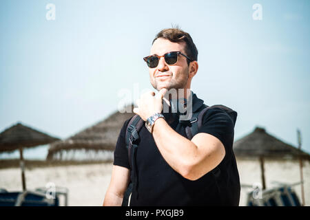 Fashionable man wearing sunglasses, headphones, watch and backpack on the beach Stock Photo