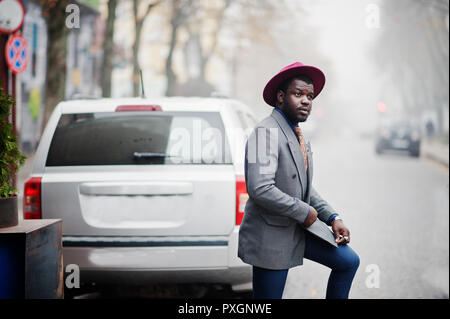 Stylish African American man model in gray jacket tie and red hat posed against silver car suv. Stock Photo