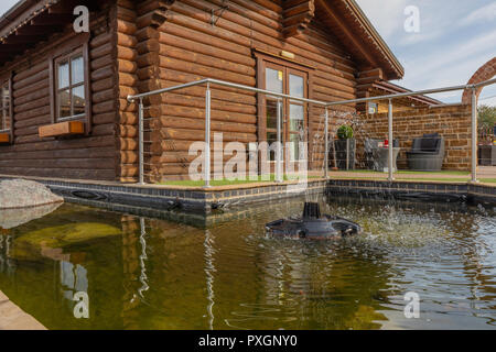 An image of a log cabin situated at the side of a fishpond with a bubbling fountain. Stock Photo