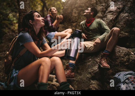 Woman sitting on rocks holding thermos with friends sitting at the back in forest. Group of friends taking a break while hiking in nature. Stock Photo