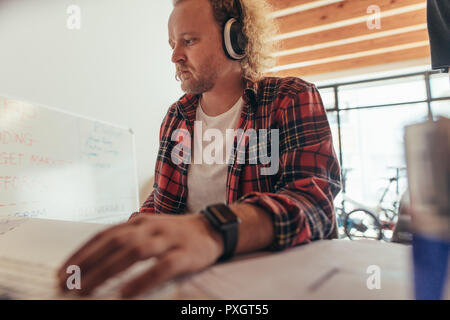 Man wearing headphones sitting at a table and reading documents. Technical support guy working in a small office. Stock Photo
