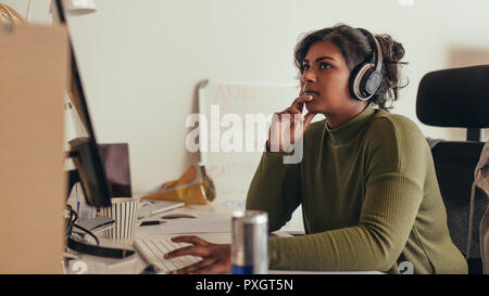 Young woman working on computer in tech startup office. Female programmer sitting at desk and looking away thinking. Stock Photo