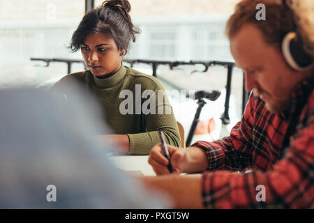 Young woman working on laptop with male colleague sitting in front writing some notes. Computer programmers working at tech startup. Stock Photo