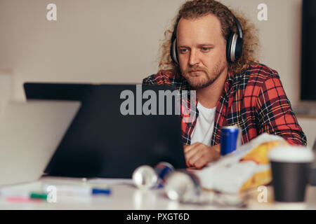 Man busy coding on laptop. Male computer programmer wearing headphones working on laptop at tech startup. Stock Photo