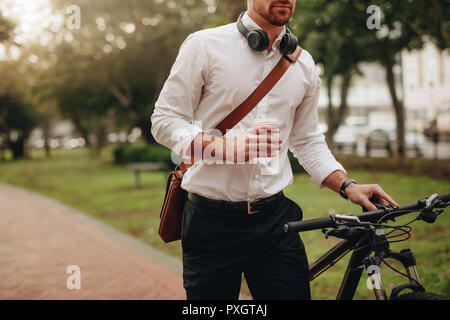 Businessman holding a coffee cup and walking to office taking his bicycle along. Man enjoying coffee while walking to work along with his bike. Stock Photo