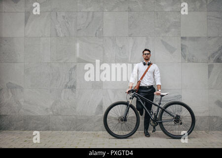 Businessman standing against a building wall in the street holding his bike. Man in formal clothes wearing office bag and headphones around the neck s Stock Photo