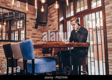 Successful bearded business man writing notary during mobile phone conversation with client, sitting at table with open laptop computer in cafe. Male  Stock Photo