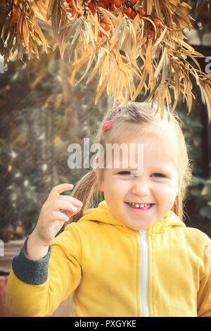 Little girl baby in yellow jumpsuit suit with blond hair gathers and bites off eats seasonal sea-buckthorn berries. Harvesting in autumn garden Stock Photo