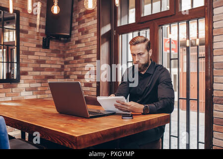 Bearded male entrepreneur makes financial report and does shopping online, buys products for restaurant, studies annual figures, analyzes profits. Ent Stock Photo
