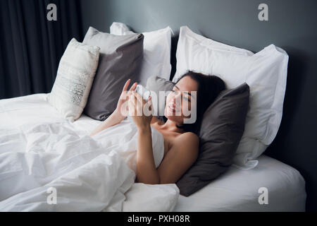 Brunette girl is reading text message from her boyfriend on mobile, while is lying in the bed. Cheerful young woman covered by blanket is watching video message on cell phone before of after sleeping. Stock Photo