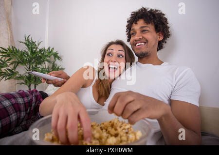 Happy beautiful couple eating popcorn while watching TV in bed and smiling Stock Photo