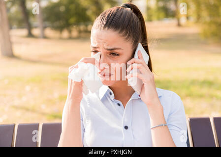 Sad young woman cries while talking on the mobile phone in the park on bench Stock Photo