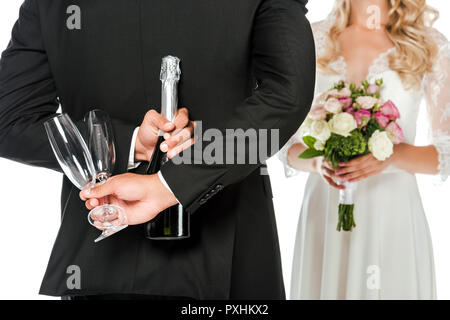 cropped shot of groom holding champagne bottle and glasses behind back while bride standing in front of him isolated on white Stock Photo