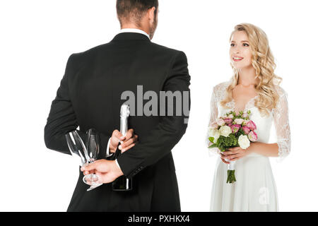rear view of groom holding champagne bottle and glasses behind back while bride standing in front of him isolated on white Stock Photo