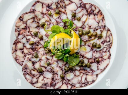 OCTOPUS CARPACCIO. Seafood Raw octopus slices with olive oil, lemon and capers on white plate. Top view. Gray stone background Stock Photo