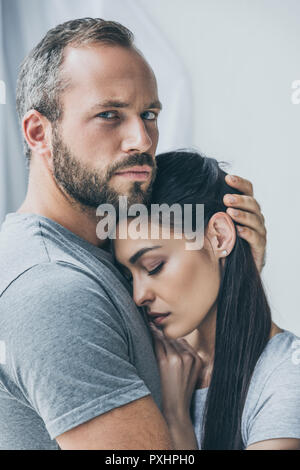 bearded man hugging upset girlfriend and looking at camera Stock Photo