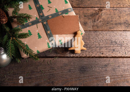 Gift wrapped in classic brown paper and green trees design, fir twigs, Christmas tree shaped gingerbread cookie and a blank tag, on a rustic table. Stock Photo