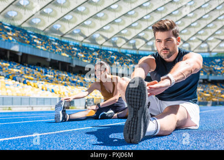 young fit couple sitting on running track and stretching at sports stadium Stock Photo