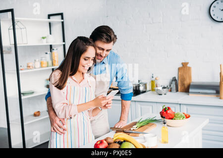 couple hugging and looking at smartphone during cooking in kitchen Stock Photo