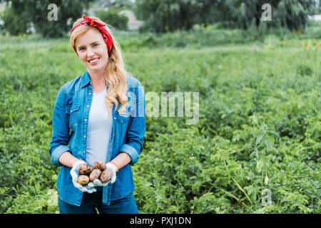 smiling attractive farmer holding ripe potatoes in field at farm and looking at camera Stock Photo