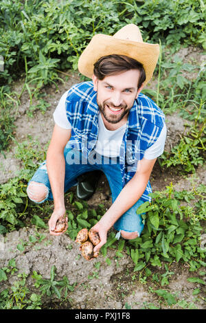 high angle view of handsome farmer squatting in field at farm and holding ripe potatoes Stock Photo