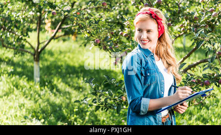smiling farmer holding clipboard in apple garden at farm and looking away Stock Photo