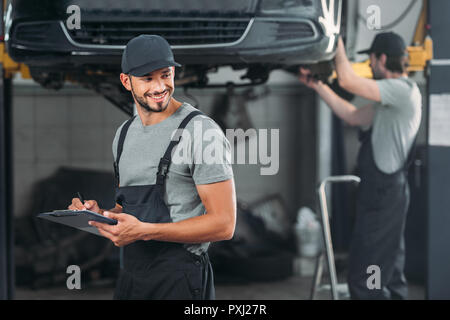 professional mechanic writing in clipboard, while colleague working in workshop behind Stock Photo