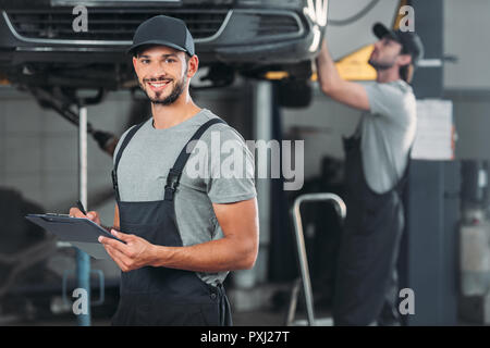 smiling mechanic writing in clipboard, while colleague working in workshop behind Stock Photo