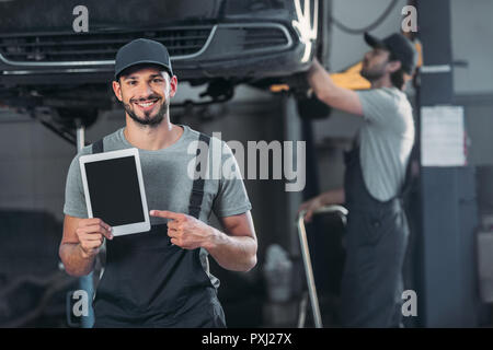 smiling mechanic showing digital tablet with blank screen, while colleague working in workshop behind Stock Photo