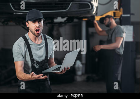 shocked mechanic using laptop, while colleague working in workshop behind Stock Photo