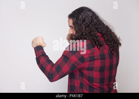 Profile side view portrait of handsome serious man with beard and black long curly hair in checkered red shirt standing with boxing gesture and attack Stock Photo