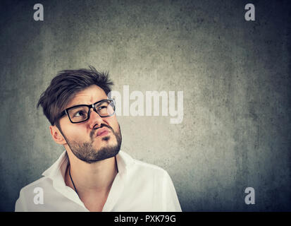 Adult bearded man in glasses and white shirt looking up in thoughts being uncertain on gray background Stock Photo
