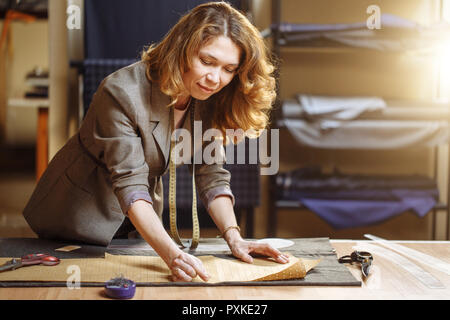 Professional adult female tailor marking cloth pattern with chalk at sewing workshop and looking at camera Stock Photo