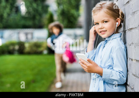 cute little child in earphones using smartphone and smiling at camera Stock Photo