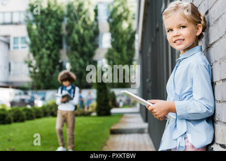 cute little schoolgirl using digital tablet and smiling at camera while leaning at brick wall Stock Photo