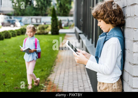 side view of schoolboy using digital tablet and schoolgirl holding book outside Stock Photo