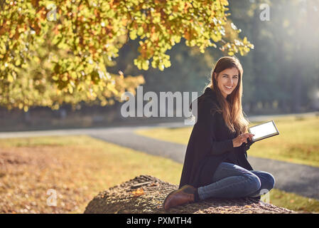 Pretty young woman relaxing in an autumn park reading on her tablet pc and smiling happily at the camera Stock Photo