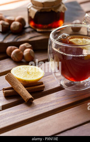 Still life composition. Tea in glass cup with lemon and vanilla pod at wood background. Hazelnut and jam in jar at wood board behind. Stock Photo