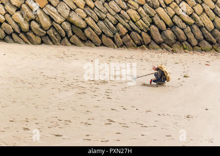 Man wearing a backpack kneeling on a sandy beach, using a metal detector on Sunny Sands beach Folkestone, Kent. UK Stock Photo