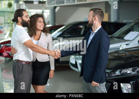 Couple making deal with car dealer, buying automobile. Beautiful smiling woman holding hand round man's waist. Bearded man shaking hands with manager and looking at each other. Stock Photo