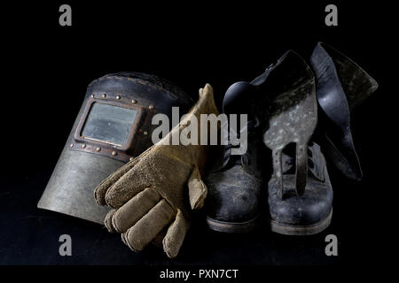 An old mask a protective suit for a welder. Occupational health and safety clothing for production workers on a workshop table. Black background. Stock Photo