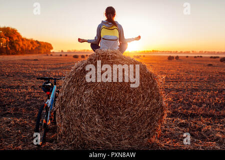 Young bicyclist meditates sitting on haystack after a ride. Woman having rest practicing yoga in autumn field. Sport recreation concept Stock Photo